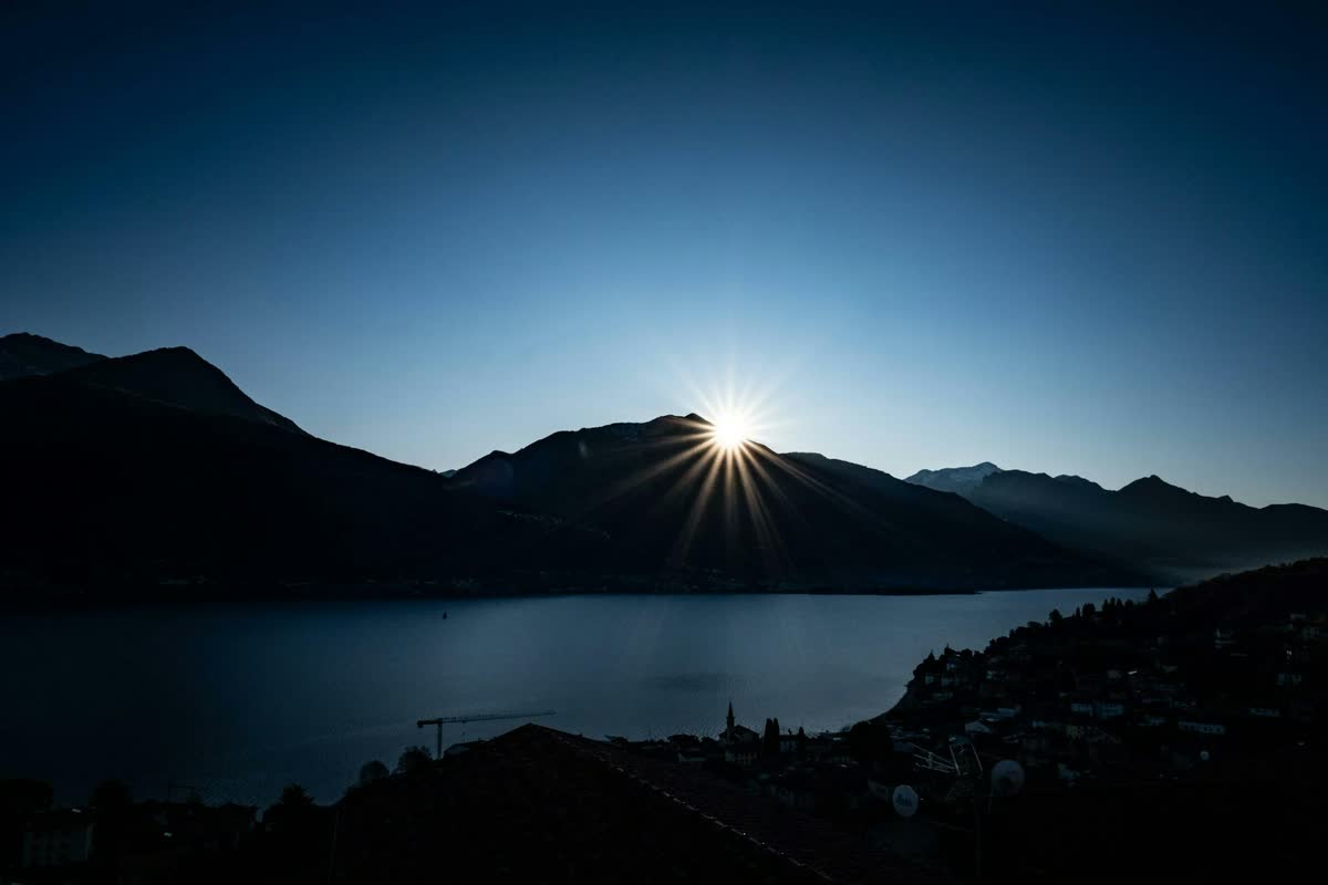 The sun breaking over a dark mountain ridge above a still lake, under a deep blue sky